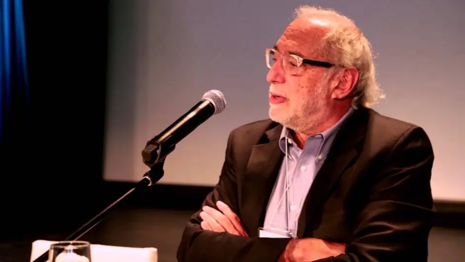 A seated man with glasses speaking into a microphone